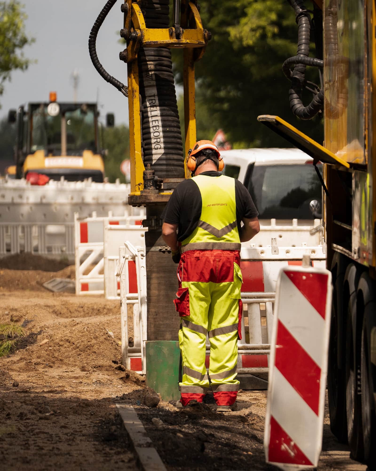 Arbeiter in gelb-roter Warnkleidung mit Gehörschutz steht von hinten gesehen an einem Bohrgerät, daneben rot-weiße Baken